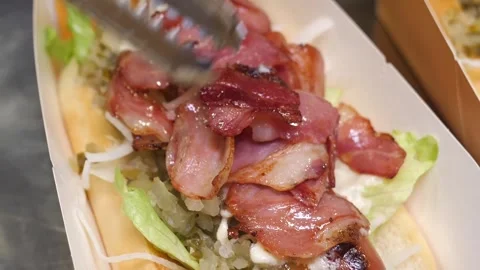 Chef placing crispy grilled bacon strips on top of gourmet hot dogs with tongs Stock Footage 324710975