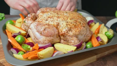 A chef is placing a whole raw marinated chicken into an oven pan with vegetables Stock Footage 145116295