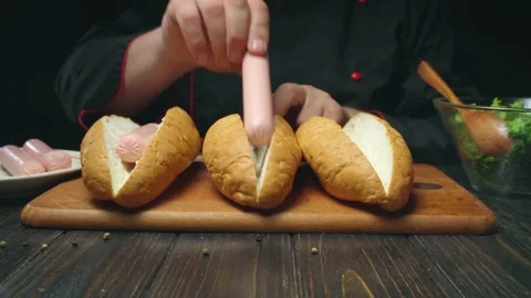 Chef prepares hot dogs by placing sausages into sliced bread rolls on a wooden Stock Footage 337374473