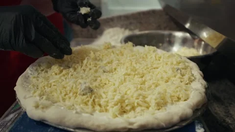 Chef in red apron prepares pizza by adding shredded cheese to dough on a blue Stock Footage 332736626