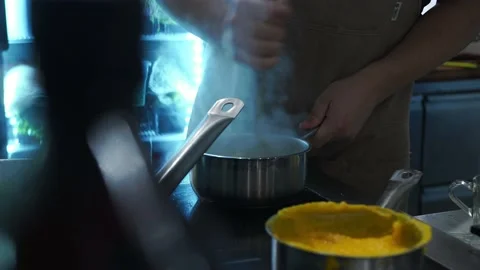 A chef in a restaurant kitchen kneads a steaming dish in a saucepan with a spoon Stock Footage 141171542
