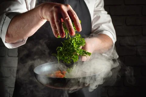 A chef is seen in a kitchen preparing a dish. They are adding fresh parsley t Stock Photos