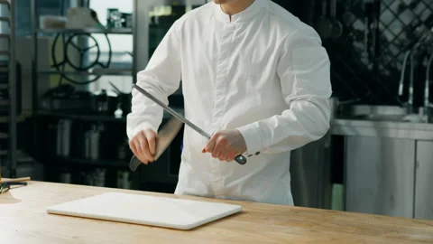 A chef is sharpening a knife at a wooden table in a professional kitchen Stock Footage 234556858