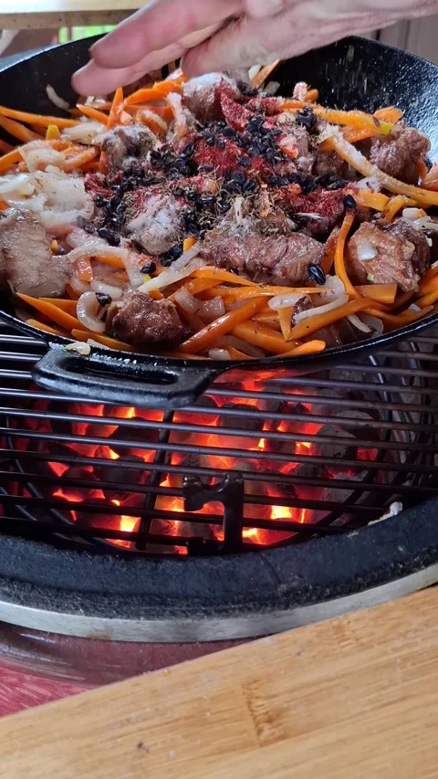 Chef sprinkling spice on meat and carrot in cast iron pan over glowing coal. Stock Footage 334623065