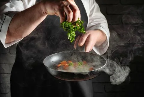 A chef stands in a kitchen, adding fresh herbs to a pan with vegetables. Th.. Stock Photos