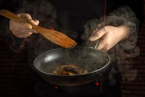 A chef stands in a kitchen, using a wooden spoon to turn a steak in a frying  Stock Photos