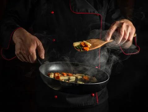 A chef is stirring chopped vegetables in a pan over a stove. The kitchen is.. Stock Photos