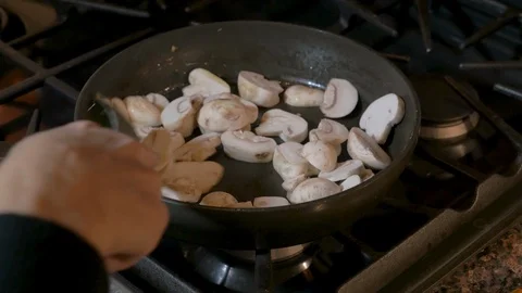 Chef stirring white sliced mushrooms in a frying pan on a stove Видео 90661153