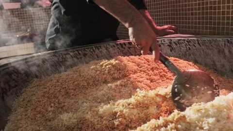 The chef stirs steaming rice in a large pilaf cauldron with a slotted spoon Stock Footage 310383750