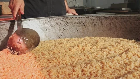 The chef stirs steaming rice in a large pilaf cauldron with a slotted spoon Stock Footage 339988535