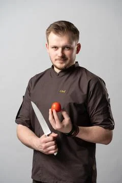 A chef in a uniform jacket shows off a professional chef's knife and a tomato Stock Photos