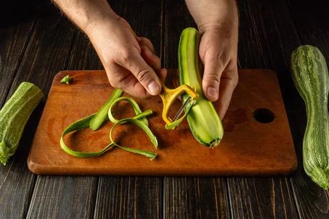 A chef uses a vegetable peeler to remove the skin from a zucchini on a wood.. Stock Photos