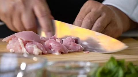A chef using a kitchen knife slices uncooked chicken meat on a chopping boa.. Stock Photos
