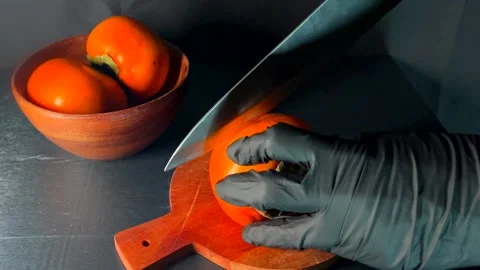Chef wearing black gloves prepares to slice ripe persimmon with large kitchen Video stock 325956599