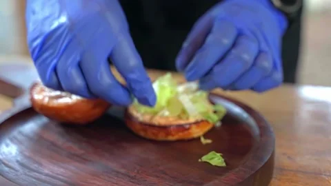 A chef wearing blue gloves puts lettuce leaves in a hamburger, slow motion video Stock Footage 228685043