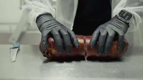 Chef in white coat and gloves slices smoked sausage on stainless steel table, Vídeo Stock 332724209