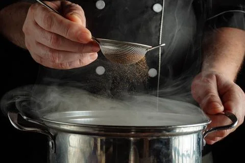 A chef works in a kitchen, holding a small strainer over a pot. The pot relea Stock Photos