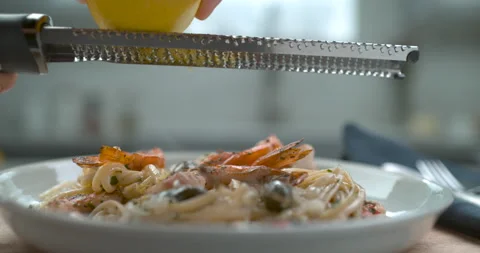 A chef's hand grates a lemon over a white plate of shrimp and olive linguini in Video stock 199456365