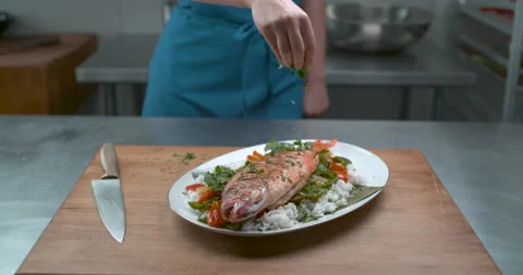 A chef's hand sprinkles herbs onto a plate of fish, rice and salad, on a wooden Video stock 199455669