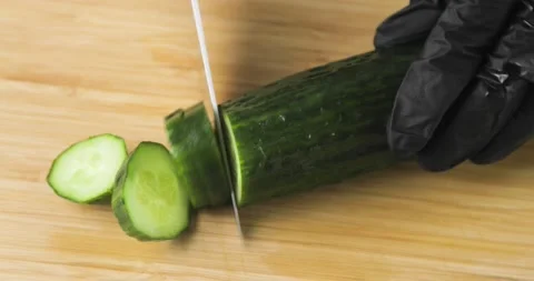 Chef's hands in black gloves cutting fresh cucumber into slices on wooden board. Stock Footage 147346632