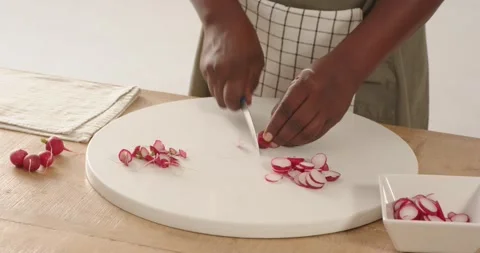 Chef's knife is slicing thin radish rounds on white cutting board on wooden Stock Footage 335269564
