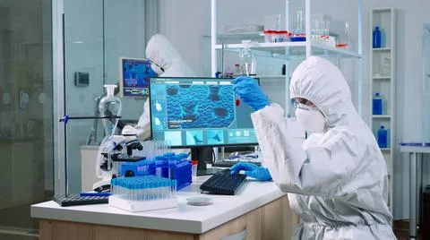Chemist in coverall checking blood samples taking notes on computer Stock Photos