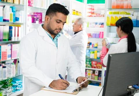 Chemist Writing On Clipboard While Colleagues Working In Pharmac Foto stock