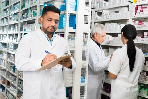 Chemist Writing On Clipboard While Colleagues Standing By Shelve Stock Photos