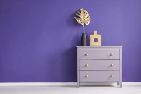 Chest of drawers with golden knobs, vases and leaf set on a purple, empty wal Stock Photos