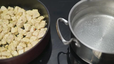 Chicken cubes are being cooked in a pan while water is being prepared in a Stock Footage 338298473
