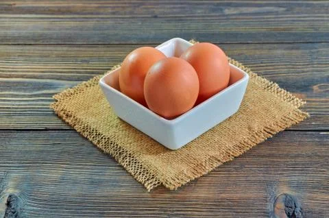 Chicken eggs in a square white clay plate on a burlap napkin, on a wooden bac Stock Photos