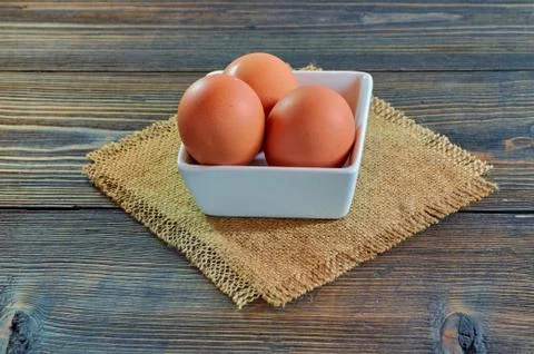 Chicken eggs in a square white clay plate on a burlap napkin, on a wooden Stock Photos