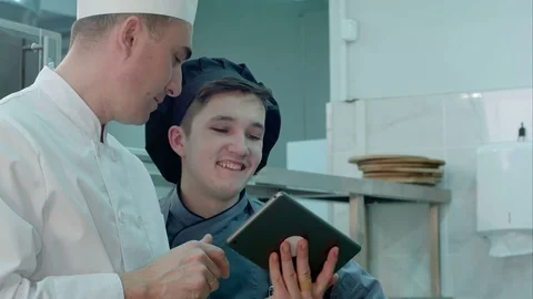 Chief chef showing cook trainee in hat something funny on digital tablet Stock Footage 72326821