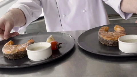 The chief puts two black dishes with fish steaks to steel table on the kitchen Stock Footage 86437216
