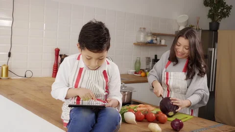 Child in apron in kitchen playing with tablet while mother is cooking vegetab Stock Footage 150664494