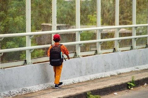 A child with a backpack and red cap walks alone on a bridge with a mesh fen.. Stock Photos