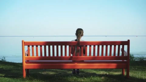 Child blond girl sitting on red bench on the shore of blue sea. Grass on beach Stock Footage