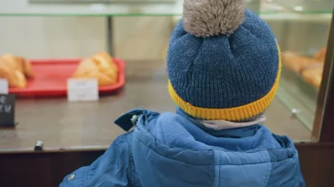 Child in blue jacket gently inquires about pastry through glass display. Little Stock Footage 345143562