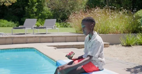Child boy cooling down in sunny backyard pool on mat, dipping feet, eating Stock Footage 337400535