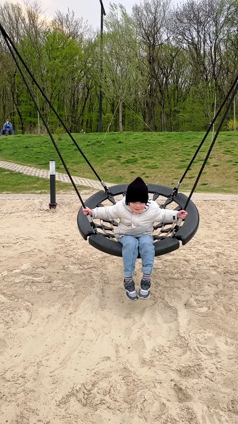 Child boy enjoys swinging on a round swing at a playground with sandy ground Stock Footage 340653013