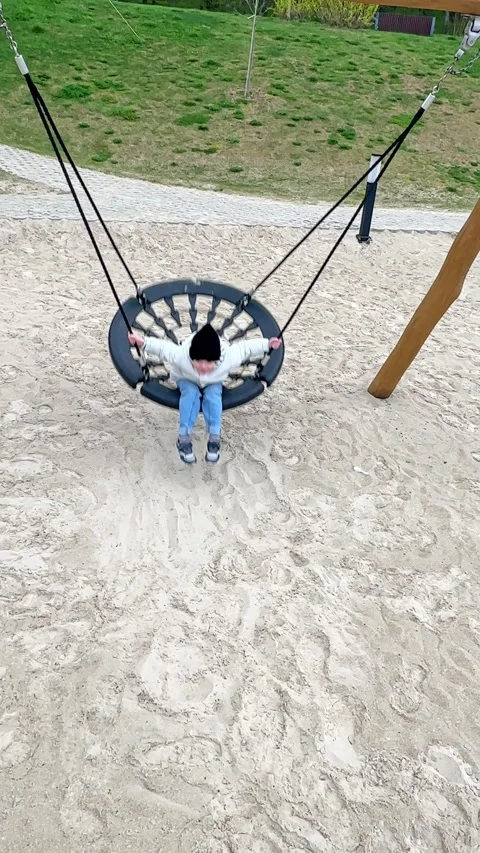 Child boy enjoys swinging on a round swing at a playground with sandy ground Stock Footage 340653190