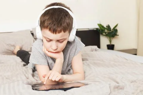 Child boy lying on bed with pc tablet with wireless headphones Stock Photos
