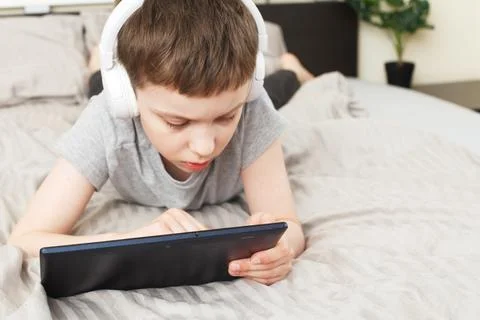 Child boy lying on bed with pc tablet with wireless headphones 写真素材