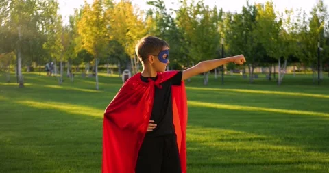 Child boy in red superhero cape stands in park. Kid wears blue mask and shows Vidéo 329866016