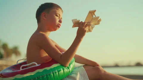 Child boy sitting alone on sandy beach on sea coast and playing with toy plane Stock Footage 317603002