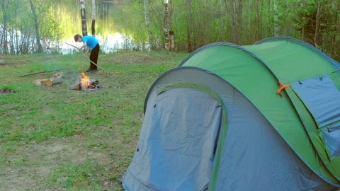 Child boy is trying to break branch for bonfire near tent on bank of lake. Stock Footage 136940575