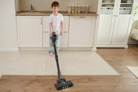 Child boy using a cordless vacuum cleaner on a kitchen floor. Small helper cl Stock Photos