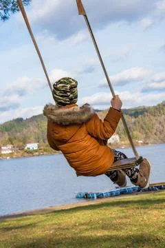 A child in a brown winter down jacket and a camouflage-colored hat swings gently Stock Photos