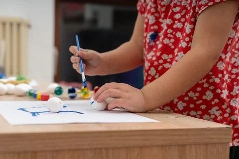 Child carefully paints easter eggs with bright blue paint on a wooden table.. 스톡 사진