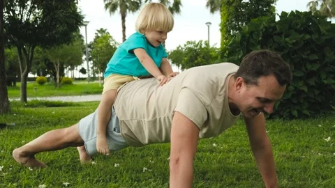 Child climbs on the father's back while father does push-ups from the floor in Stock Footage 200807582
